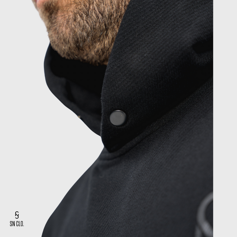 Close-up of a SN Clo. black hooded garment with a button on a white background