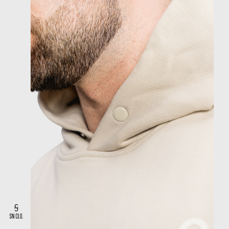Close-up of a person wearing a beige SN Clo. hoodie showing button detail with a brand logo in the corner.